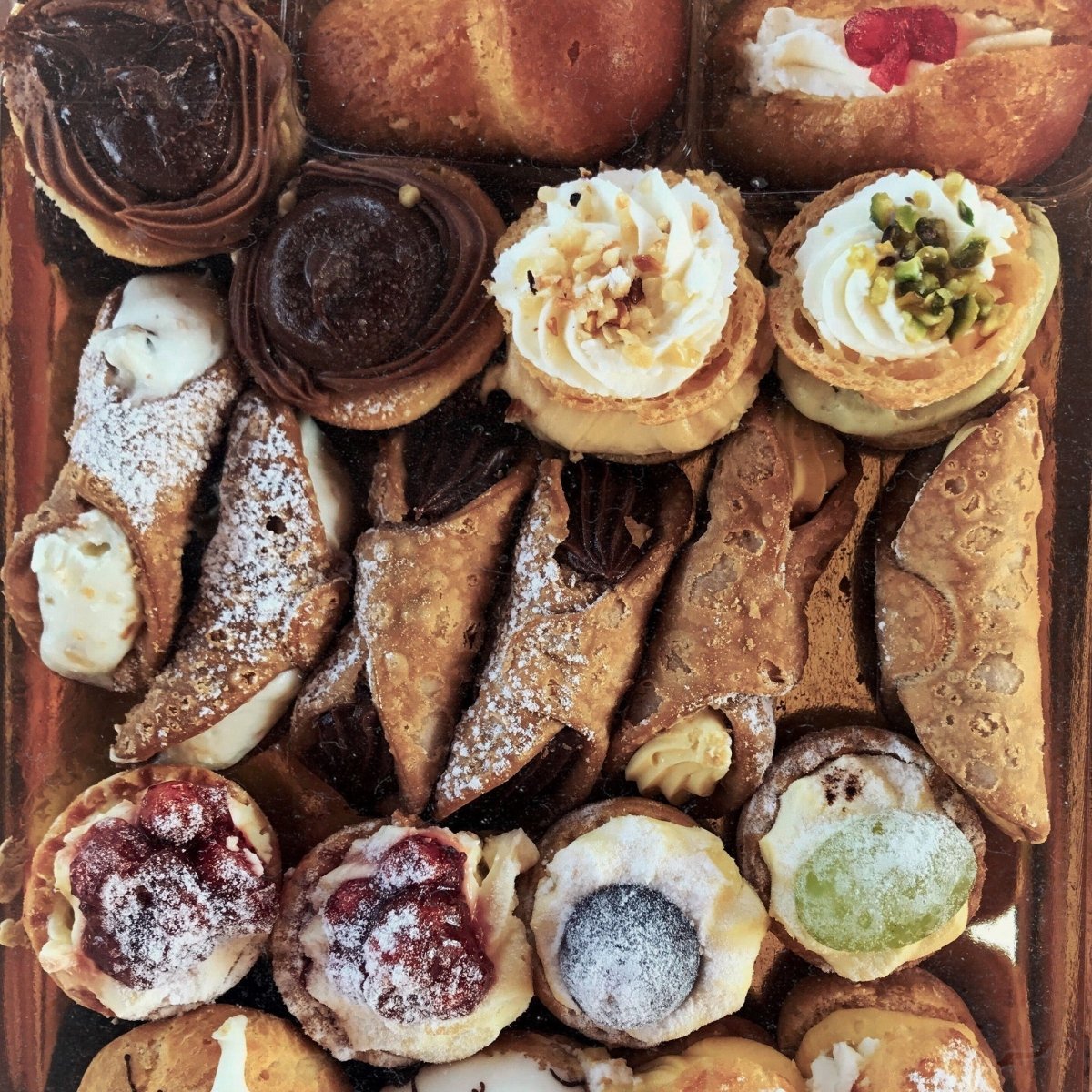 Overhead view of a tray filled with cream-filled cannoli and assorted pastries topped with chocolate, nuts, fruit, and powdered sugar.