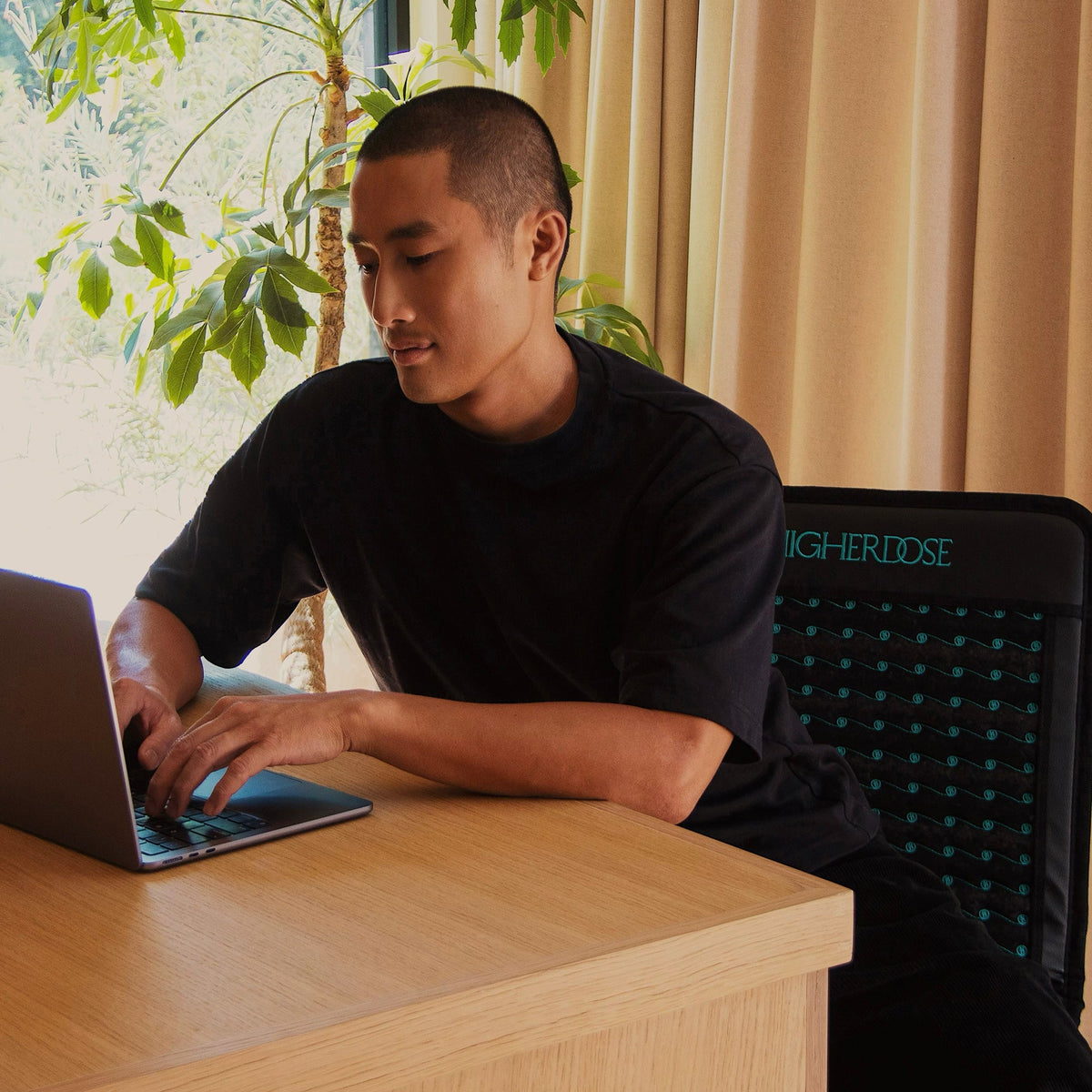 Man working on a laptop at a light wood desk with the HigherDOSE Infrared PEMF Go Mat behind him