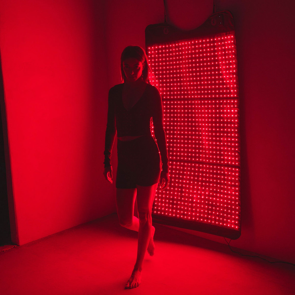 Woman standing in front of the hung HigherDOSE Full Body Red Light Mat glowing red in a dark room