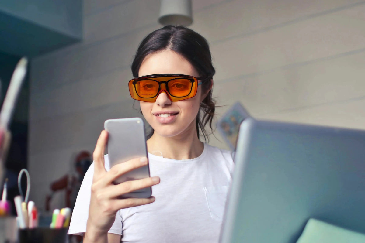 Woman looking at her phone in front of a laptop wearing Lumos Sleep Blue Blocker orange glasses