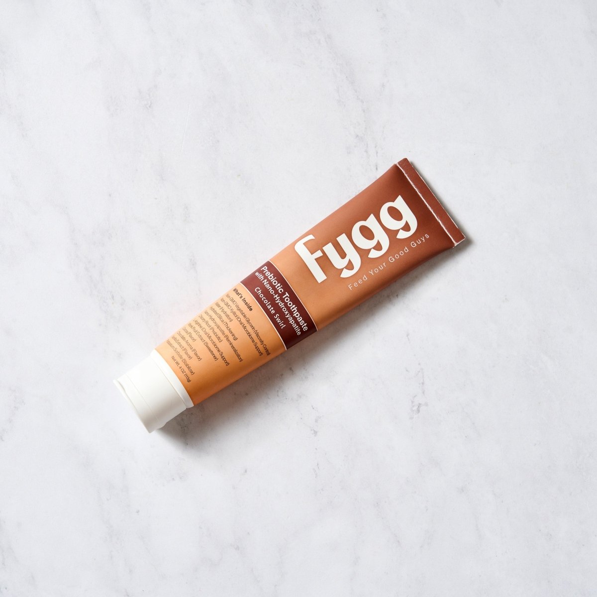 A squeeze tube of Fygg Hair & Skin Cream placed diagonally on a light gray marble surface. The tube is brown with a white cap, featuring the brand name 'Fygg' in white text.