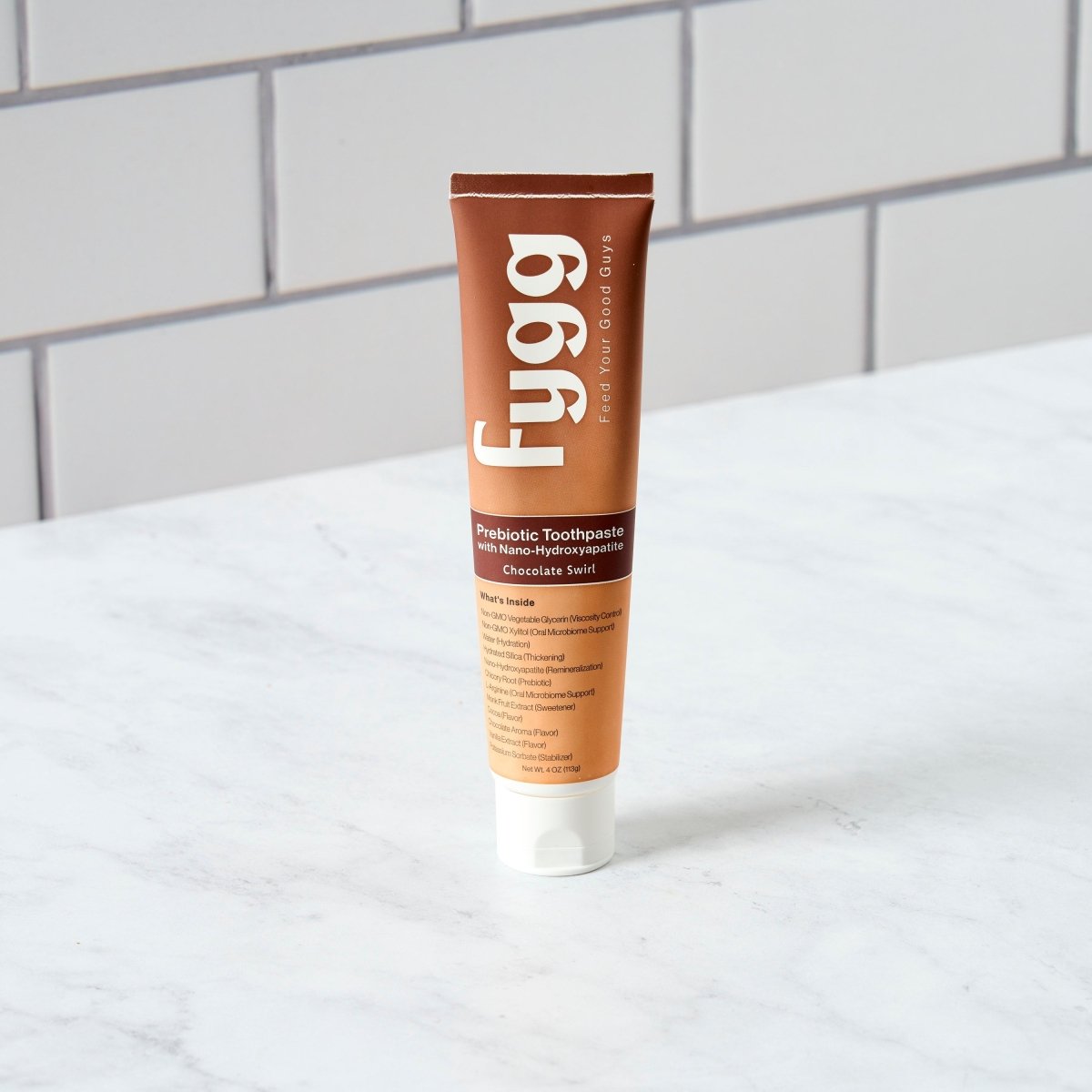 A tube of Fygg Peribiotic Toothpaste in Chocolate Scent, standing upright on a white marble countertop with a white subway tile backsplash in the background.