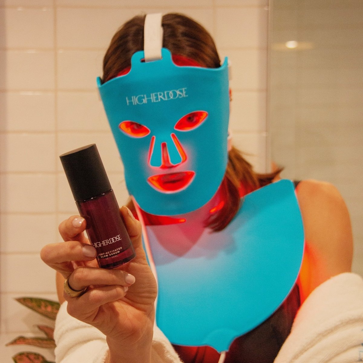 Woman in a robe wearing the HigherDOSE Red Light Face Mask and Neck Enhancer, holding the Glow Serum in front of her
