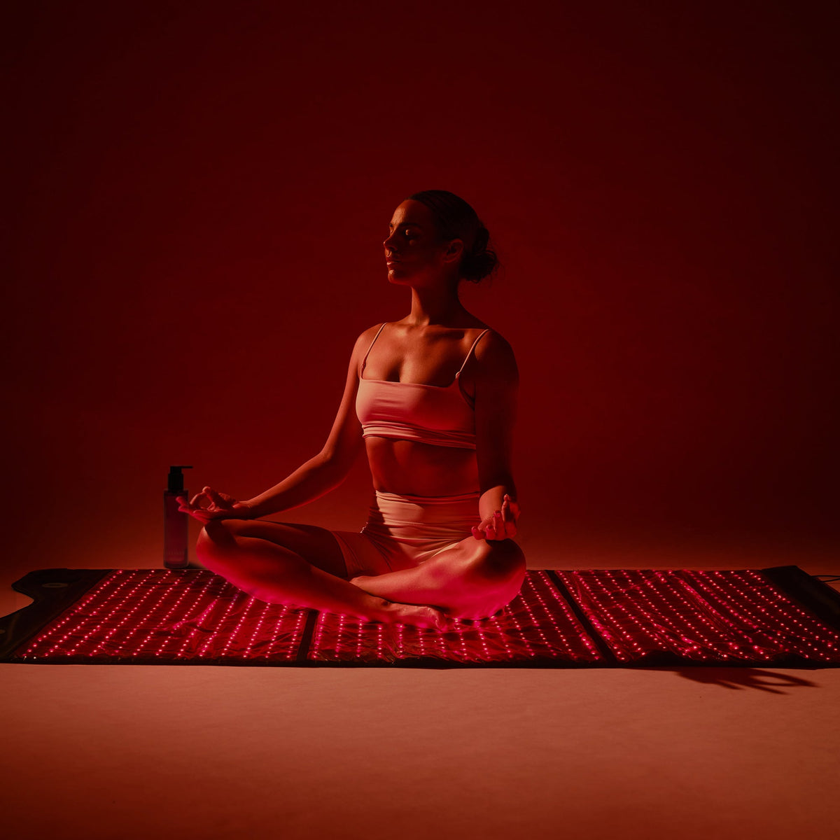 Woman meditating on the HigherDOSE Full Body Red Light Mat with a body oil bottle beside her