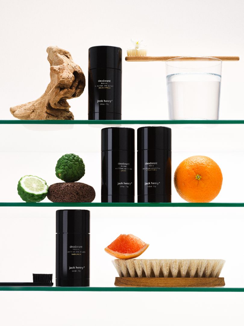 Glass bathroom shelves holding Jack Henry Deodorants, a brush, a toothbrush, citrus and driftwood
