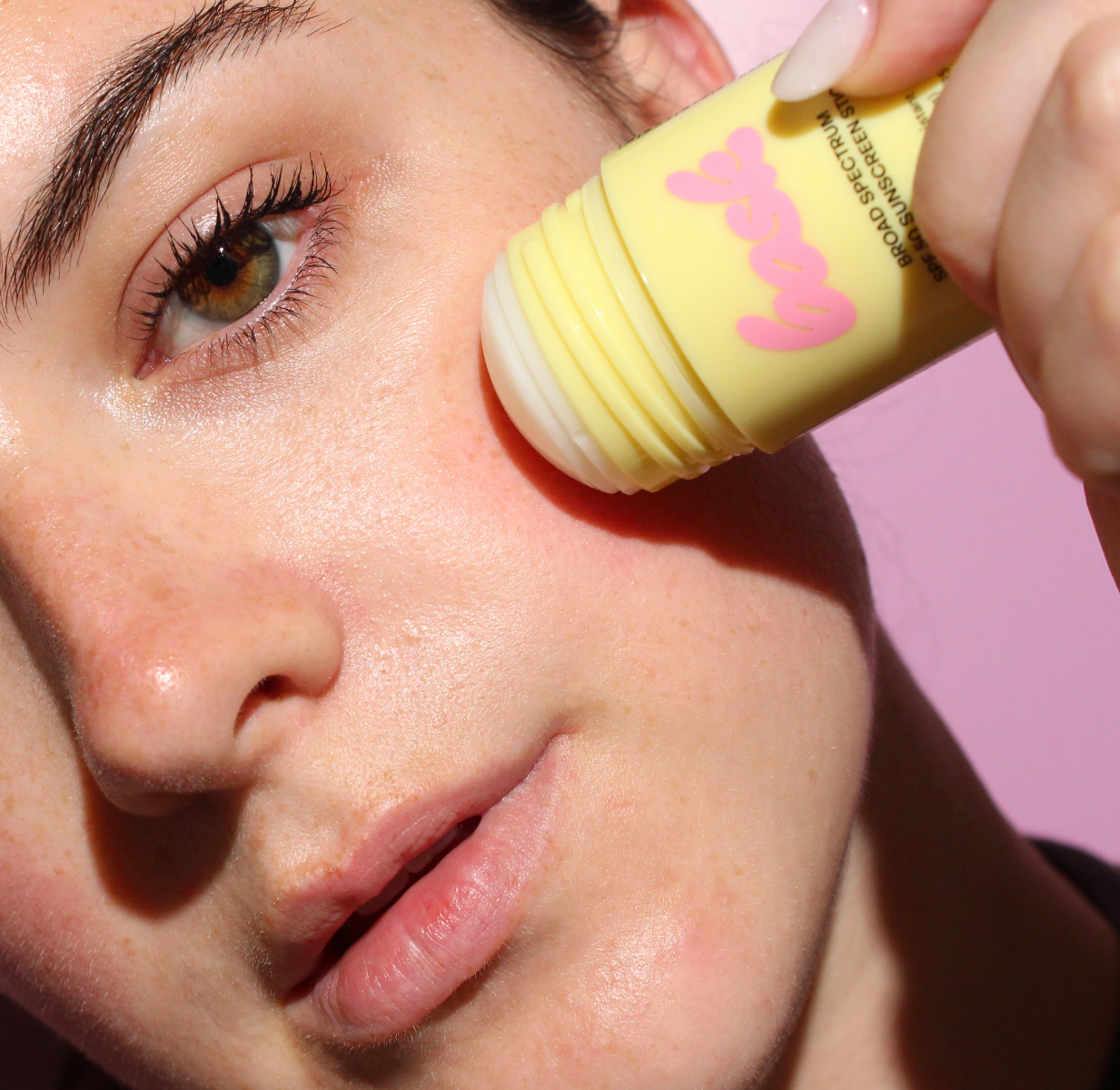 Person applying a yellow bask sunscreen to their face with a pink applicator on a pink background