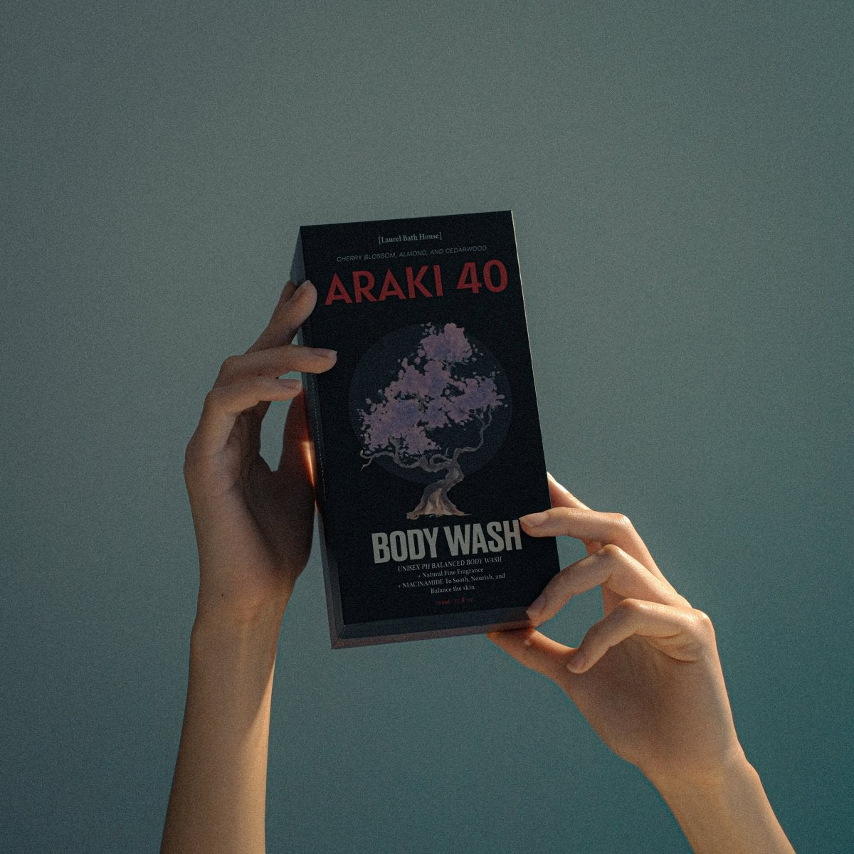 Hands holding the dark Laurel Bath House Araki 40 body wash box against a teal background
