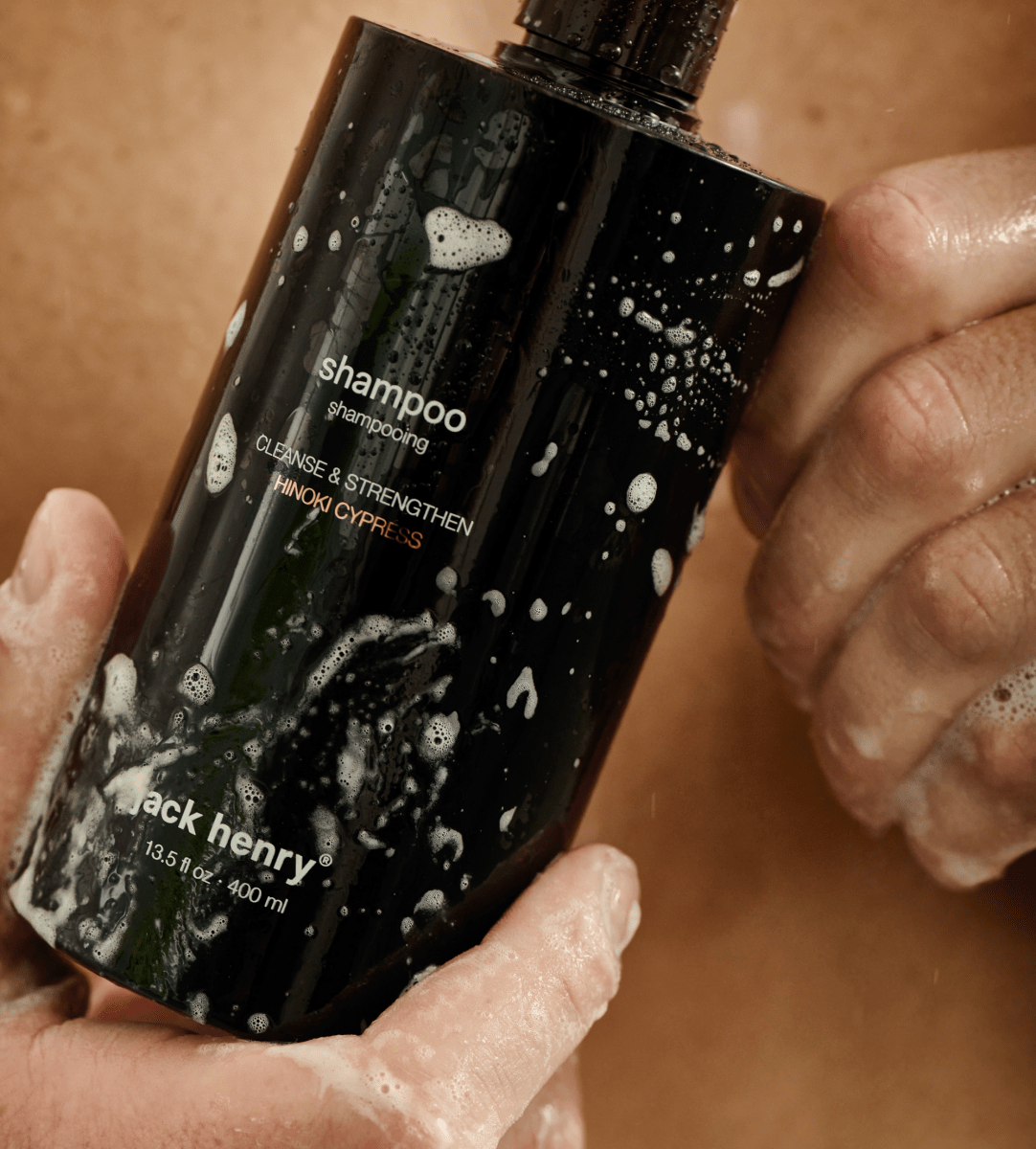 Wet hands holding a suds-covered bottle of Jack Henry Creamy Shampoo