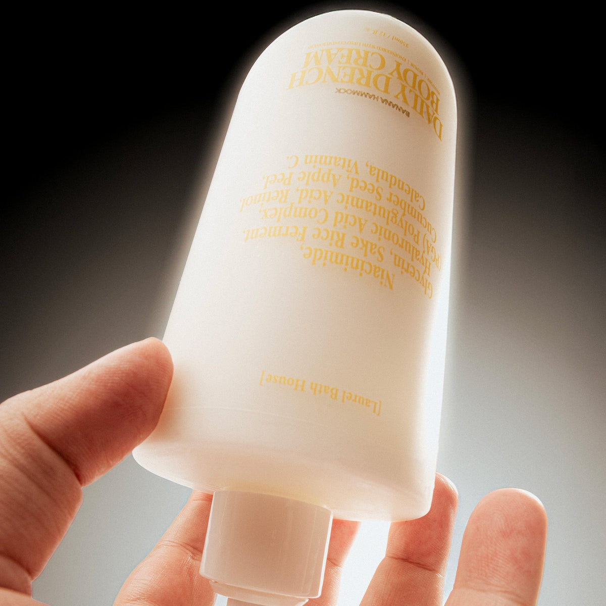 Hand holding the Banana Hammock Laurel Bath House Daily Drench Body Cream bottle upside down, lit from behind