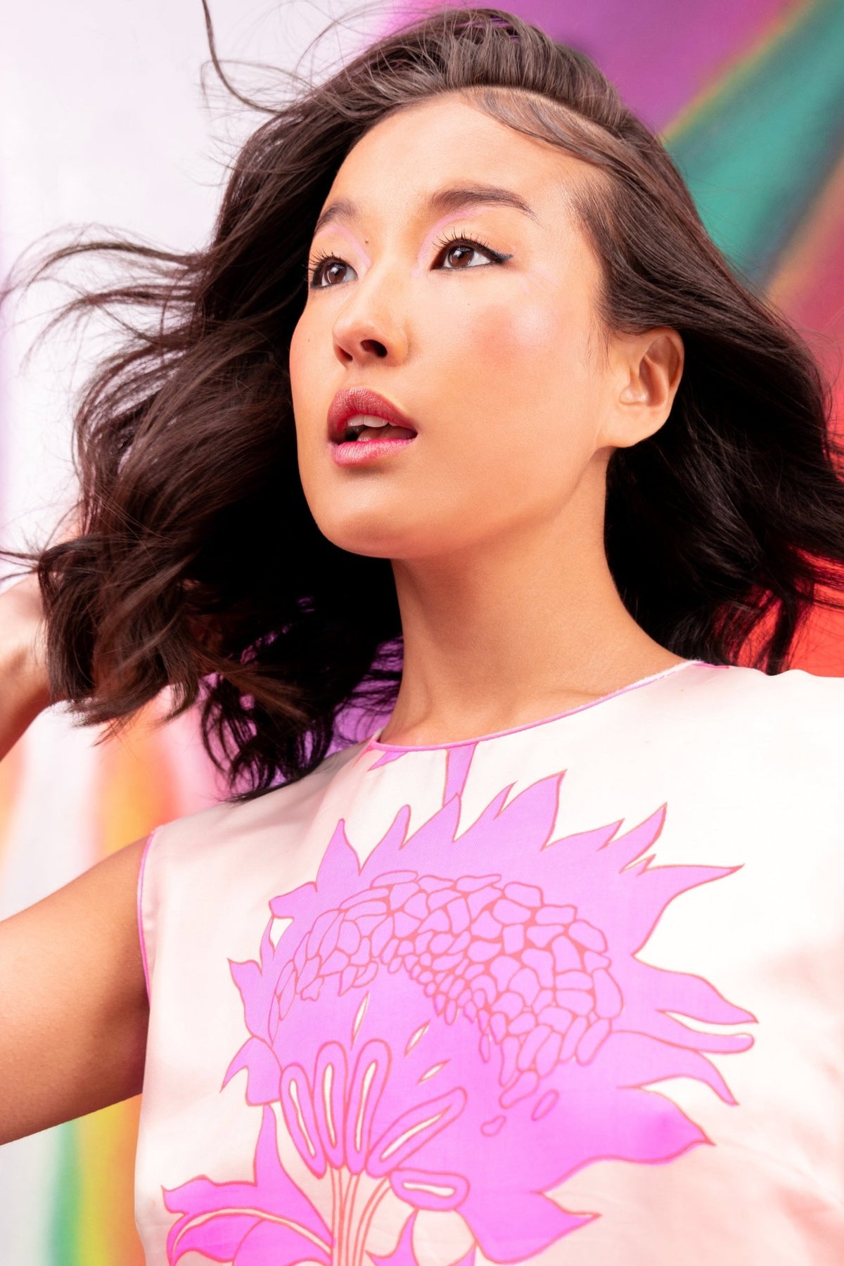 Woman with wind-blown hair and a pink floral top wearing TANAÏS Heart Chakra Mood Change Lip Stain against a bright wall