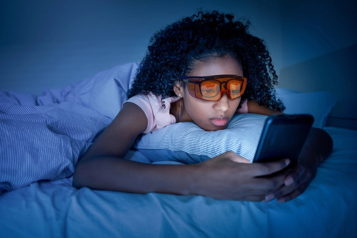 Woman lying in bed looking at her phone wearing Lumos Sleep Blue Blocker orange glasses