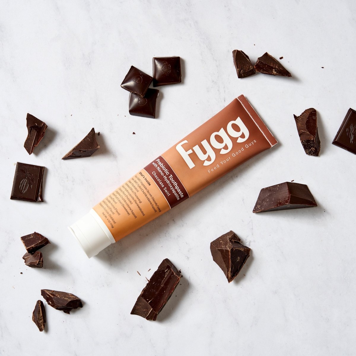 A tube of Fygg cocoa butter lip balm lies diagonally on a white marble surface, surrounded by scattered dark chocolate pieces.