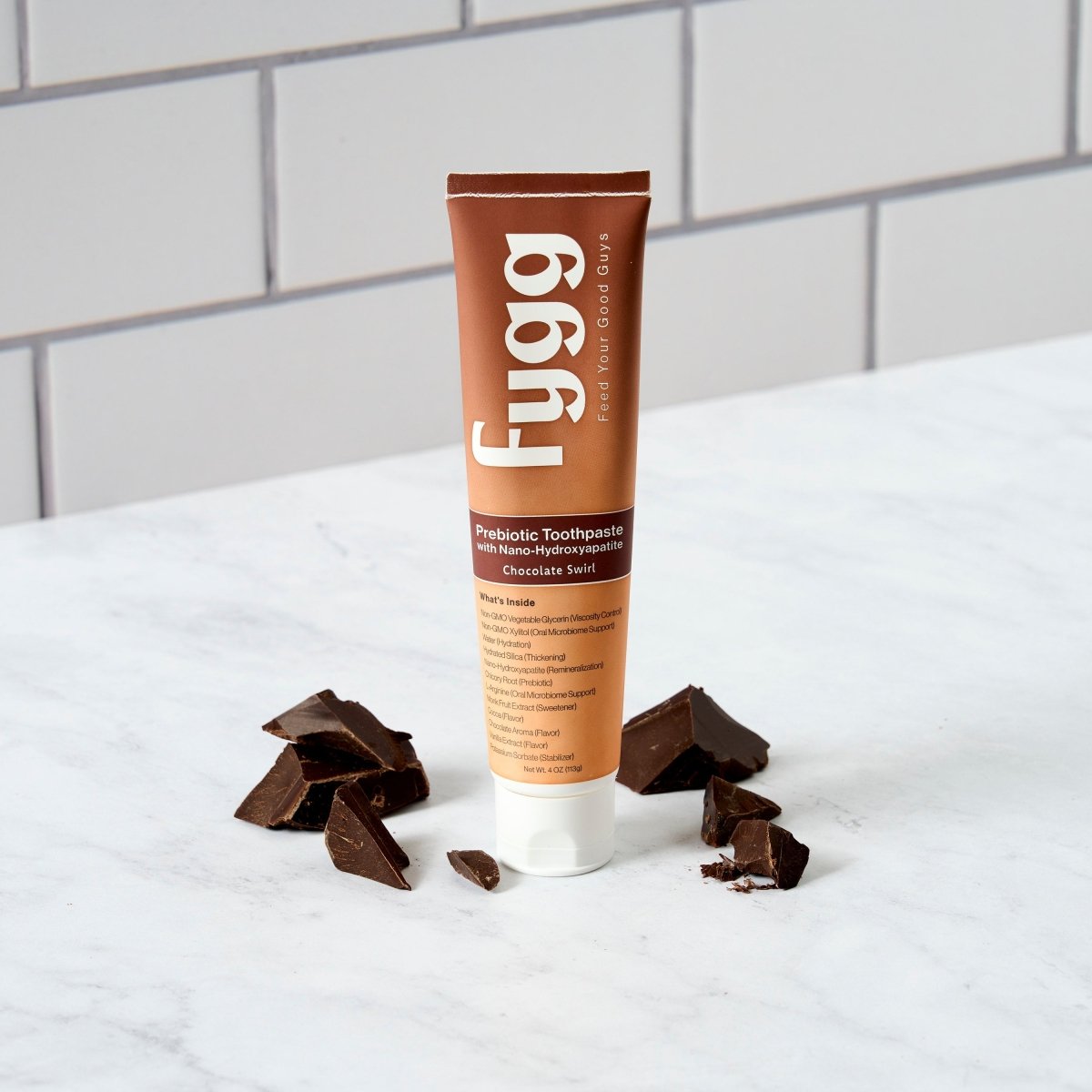 A tube of Fygg Prebiotic Toothpaste in Chocolate Sea Salt flavor, styled on a white marble surface with pieces of dark chocolate scattered around it against a white subway tile background.