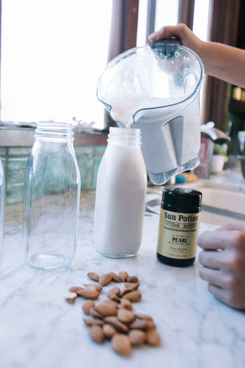 Hand pouring a milk drink from a blender beside a Sun Potion Pearl Powder jar and almonds