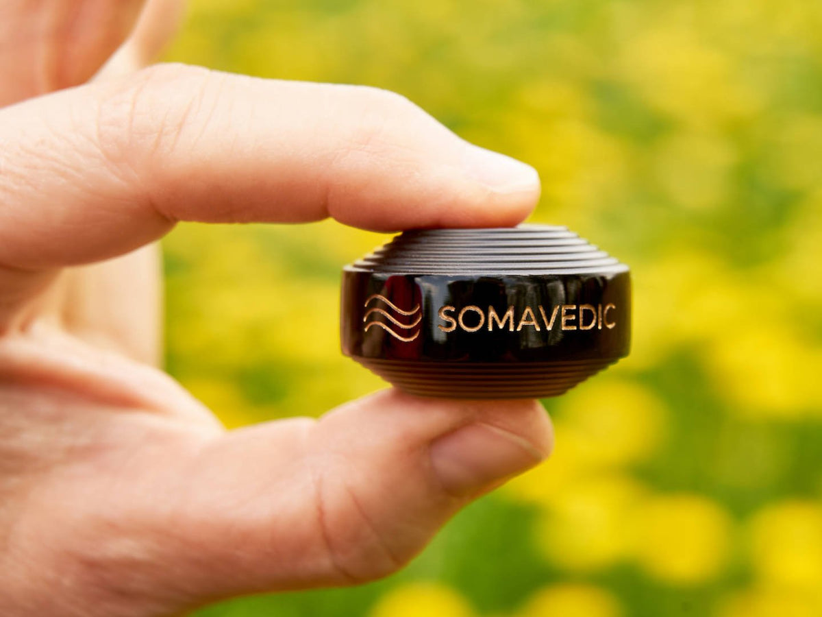 Fingers holding the Somavedic Tiny black against a yellow flower field