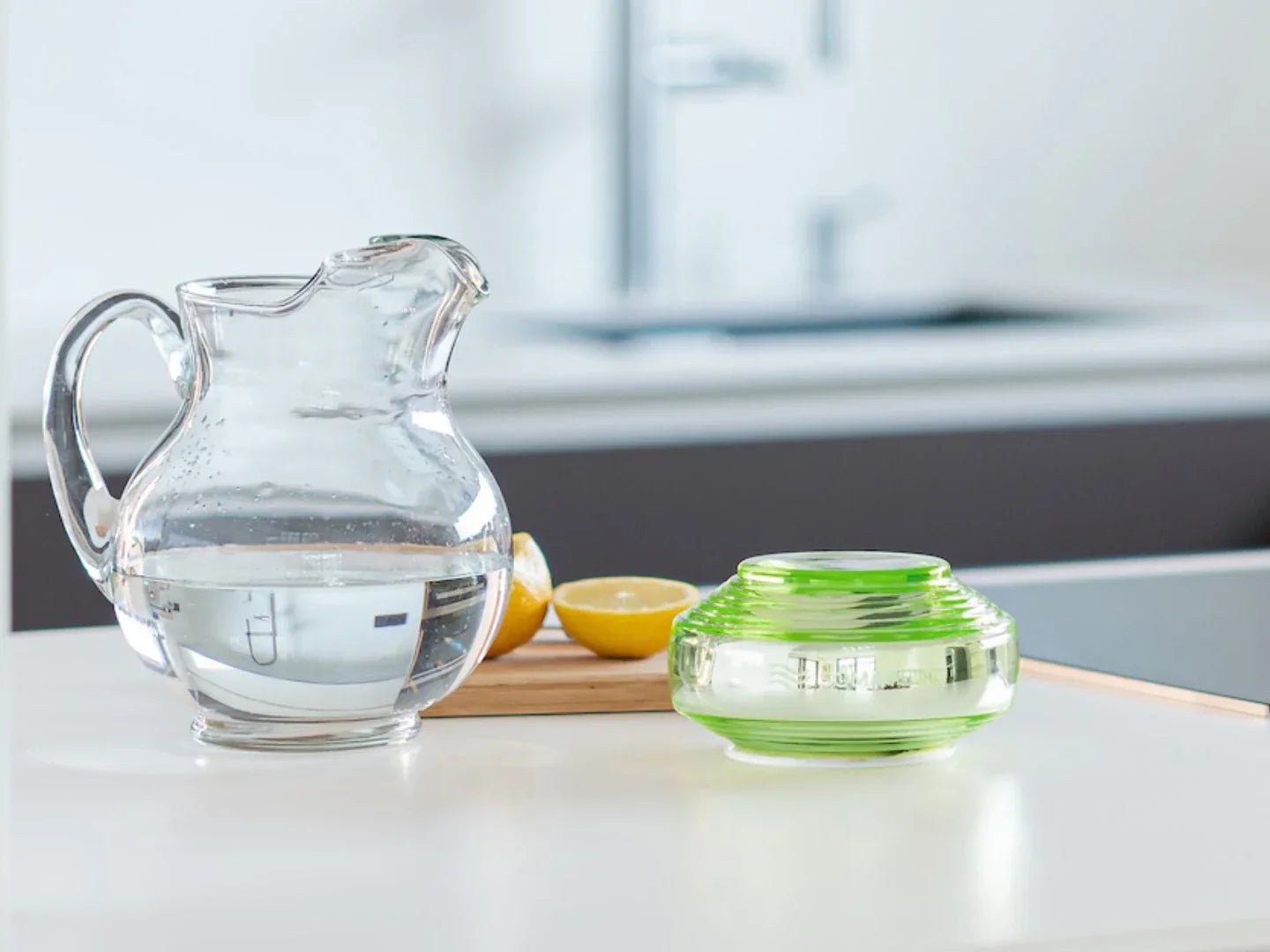 Clear glass pitcher with water on a kitchen counter with lemons and a green container.